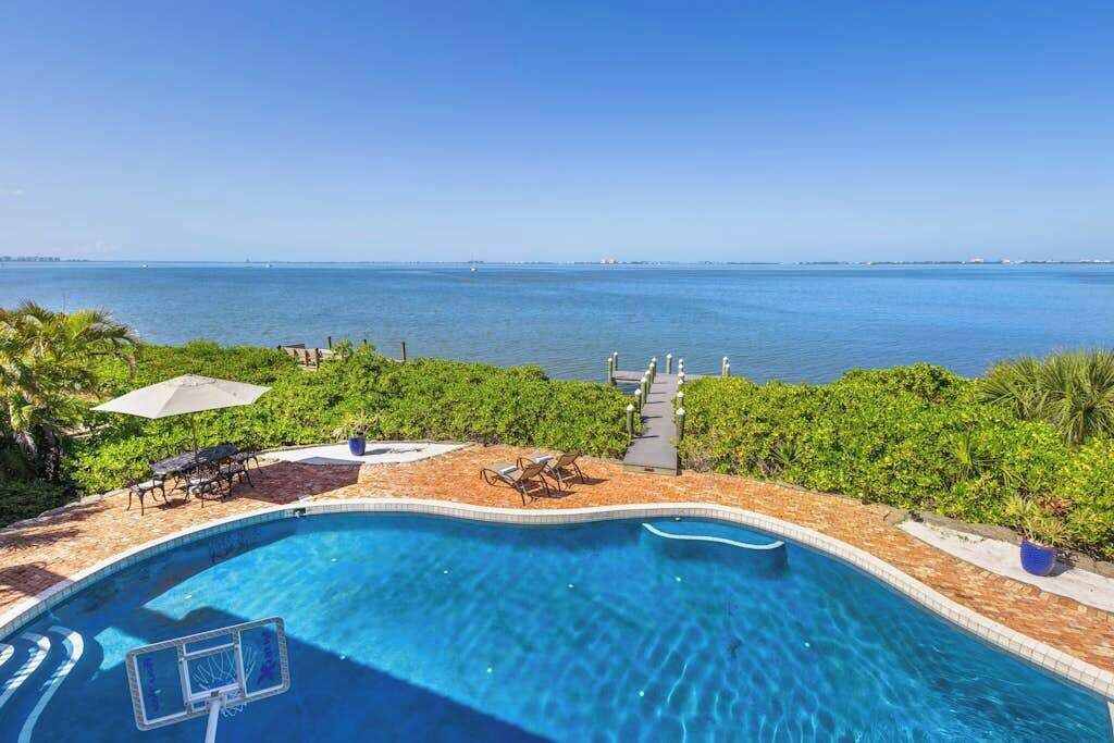 A swimming pool with a basketball hoop, surrounded by greenery and overlooking a body of water.