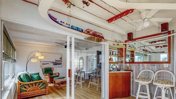 A room with a glass door leading to a dining area, a sofa, a bar area with stools, and a ceiling with exposed pipes and a fan. - Waterfront Home with  Pool, Private Washer & Dryer, and Gazebo (Bradenton)