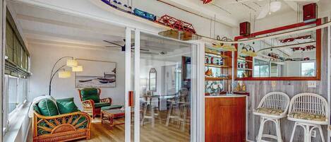 A room with a glass door leading to a dining area, a sofa, a bar area with stools, and a ceiling with exposed pipes and a fan.