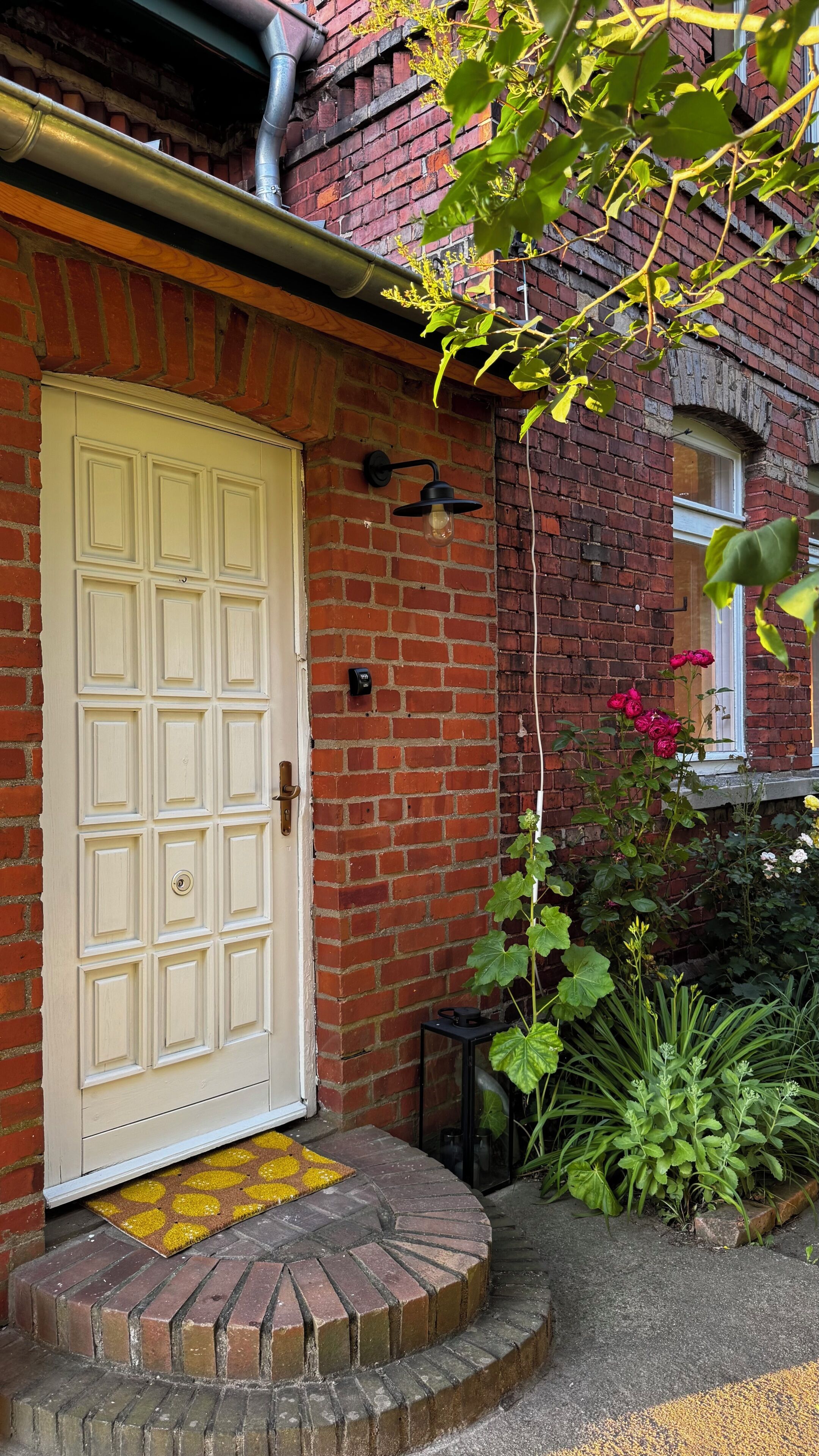A white door with a small step leading up to it, a brick wall, a black wall-mounted light fixture, and a window with a white frame.