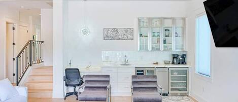 Ein modernes Wohnzimmer mit einer Couch, zwei Sesseln, einem Couchtisch, einer Küchenzeile mit Schränken, einem Kühlschrank und einem an der Wand befestigten Fernseher.