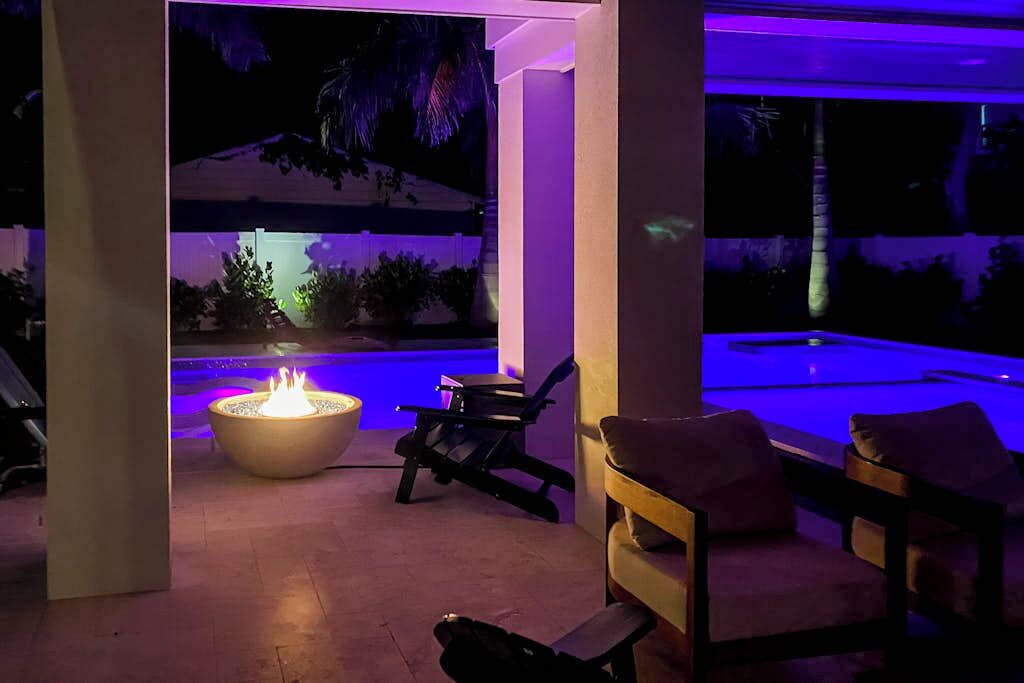 An outdoor seating area with a fire feature, a pool, and a view of a landscaped area.