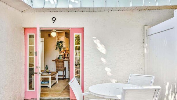 Un patio con una mesa y sillas blancas, una puerta rosa que da a un espacio interior, y el número '9' en la pared.