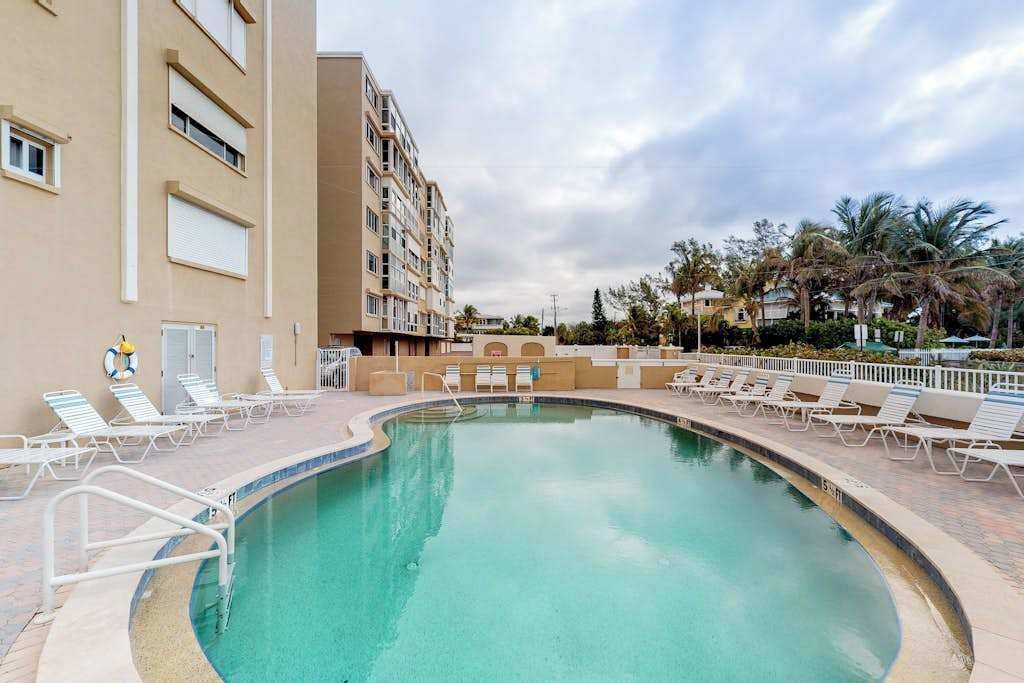 A swimming pool with lounge chairs, a building with balconies, and palm trees in the background.