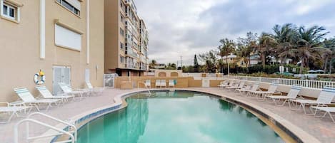 A swimming pool with lounge chairs, a building with balconies, and palm trees in the background.