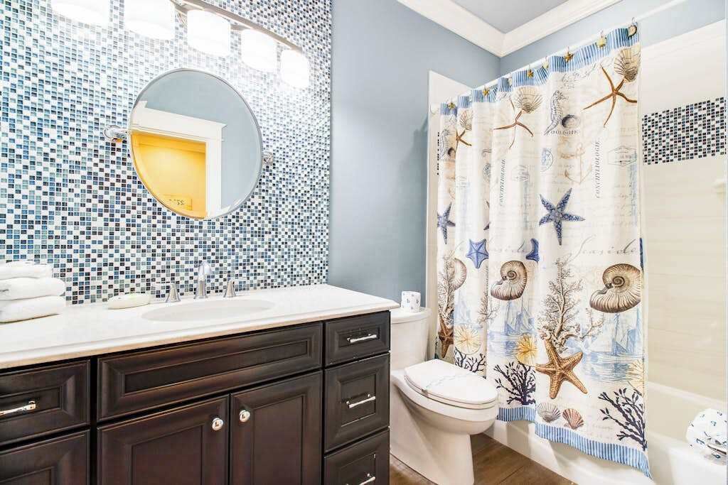 A bathroom with a mosaic tile wall, a round mirror, a dark wooden vanity, a white toilet, and a shower curtain with sea-themed designs.