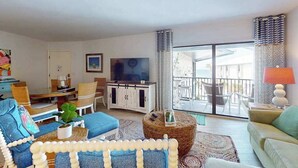 Uma sala de estar com sofá, poltrona, mesa de jantar, televisão e varanda com mobiliário de exterior.