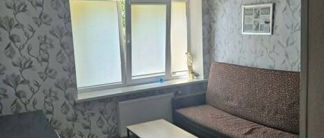 Une pièce comprenant un canapé, une petite table, un radiateur, une fenêtre à vitres dépolies et un ventilateur de plafond.