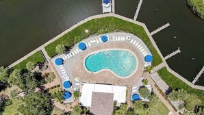 Luchtbeeld van een resort met een centraal zwembad, omringd door ligbedden en parasols, met een steiger en boten op de achtergrond.