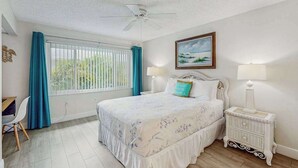 Une chambre à coucher comprenant un lit, un bureau, une chaise, une fenêtre avec des rideaux, un ventilateur de plafond et un tableau accroché au mur.