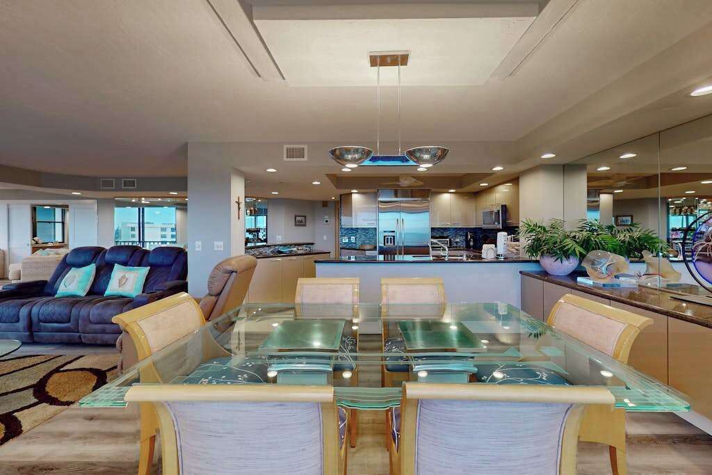 A modern living room with a glass dining table, leather sofa, and a kitchen in the background.