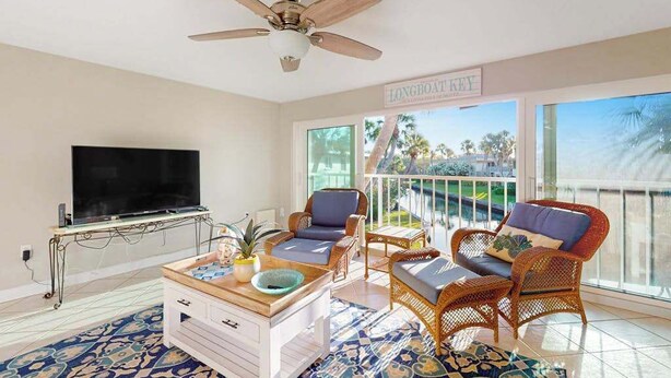 A living room with a TV, wicker furniture, a ceiling fan, and a balcony with a view. - Waterfront condo with Gulf views, pool, tennis, & deeded beach access (Longboat Key)