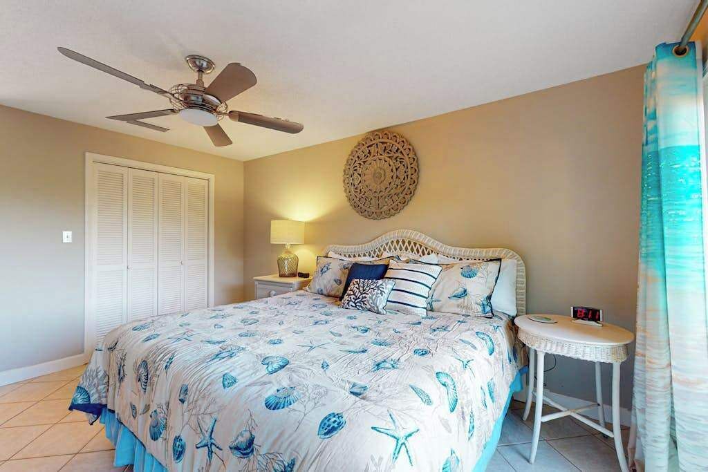 A bedroom with a bed, a ceiling fan, a bedside table with a lamp, a decorative wall piece, and a window with curtains.