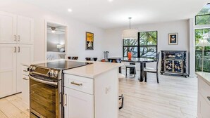 Une cuisine moderne avec des meubles blancs, un four encastré, un coin repas avec une table et des chaises, et une vue sur la verdure à l’extérieur.
