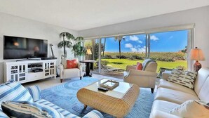A living room with a TV, sofas, a coffee table, and a large window with a view of a green landscape. - Beachfront Condo with a Beautiful, Renovated Kitchen, Shared Pool, & Tennis (Longboat Key)