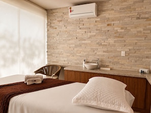 Habitación de hotel con pared de ladrillo, cama con ropa blanca, cabecera de madera, baño con lavamanos y artículos de aseo, y un equipo de aire acondicionado.