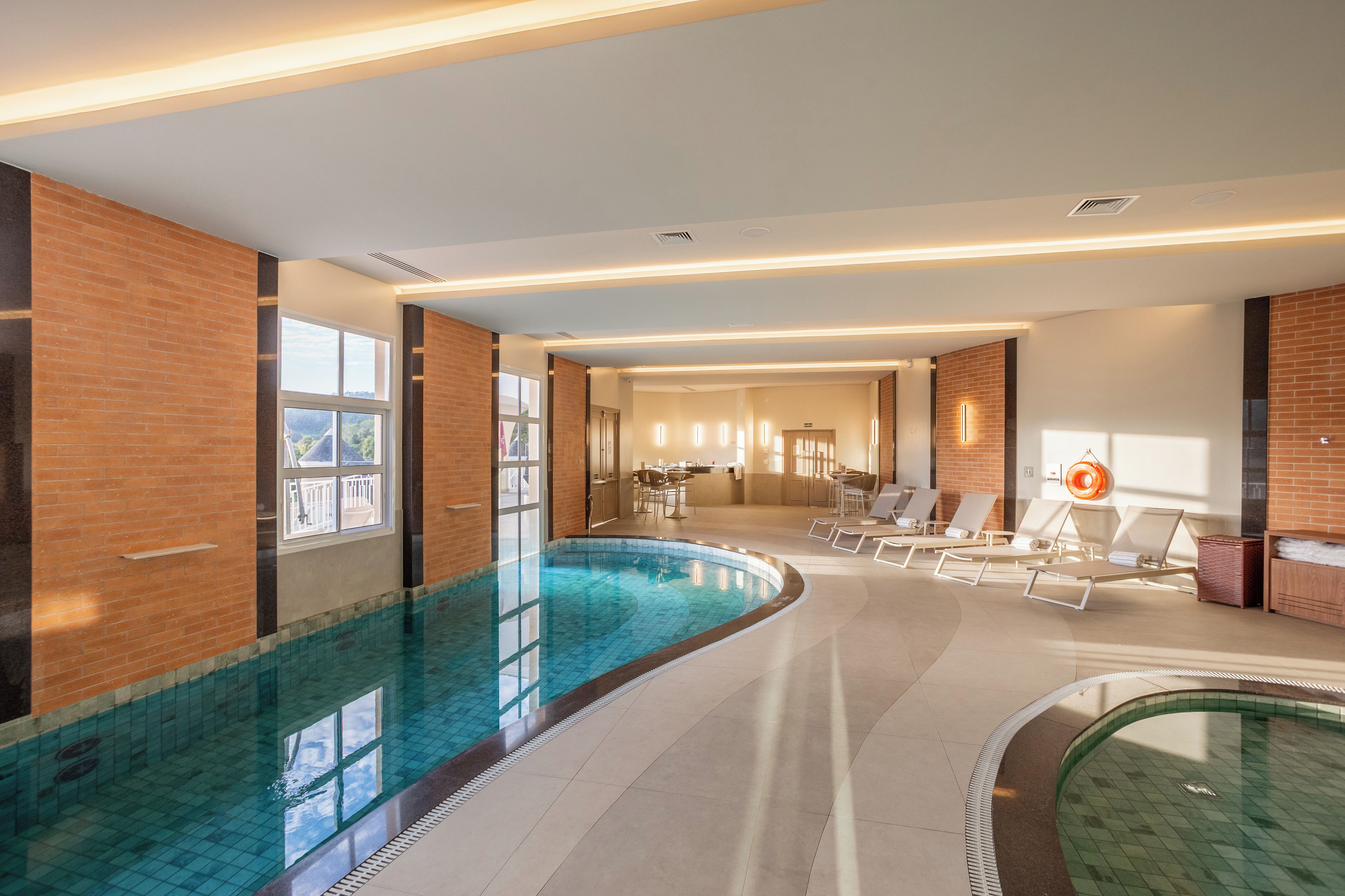 Piscina interna com piso de azulejo, área de estar com espreguiçadeiras e parede de tijolos.
