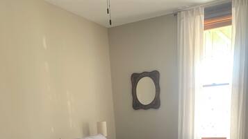 Un llit doble amb edredó gris, una petita taula de nit amb una làmpada, un mirall a la paret i una finestra amb cortines.