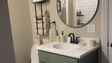 Et badeværelse med en hvid vask, et rundt spejl, et toilet og en håndklædestang med sammenrullede håndklæder.