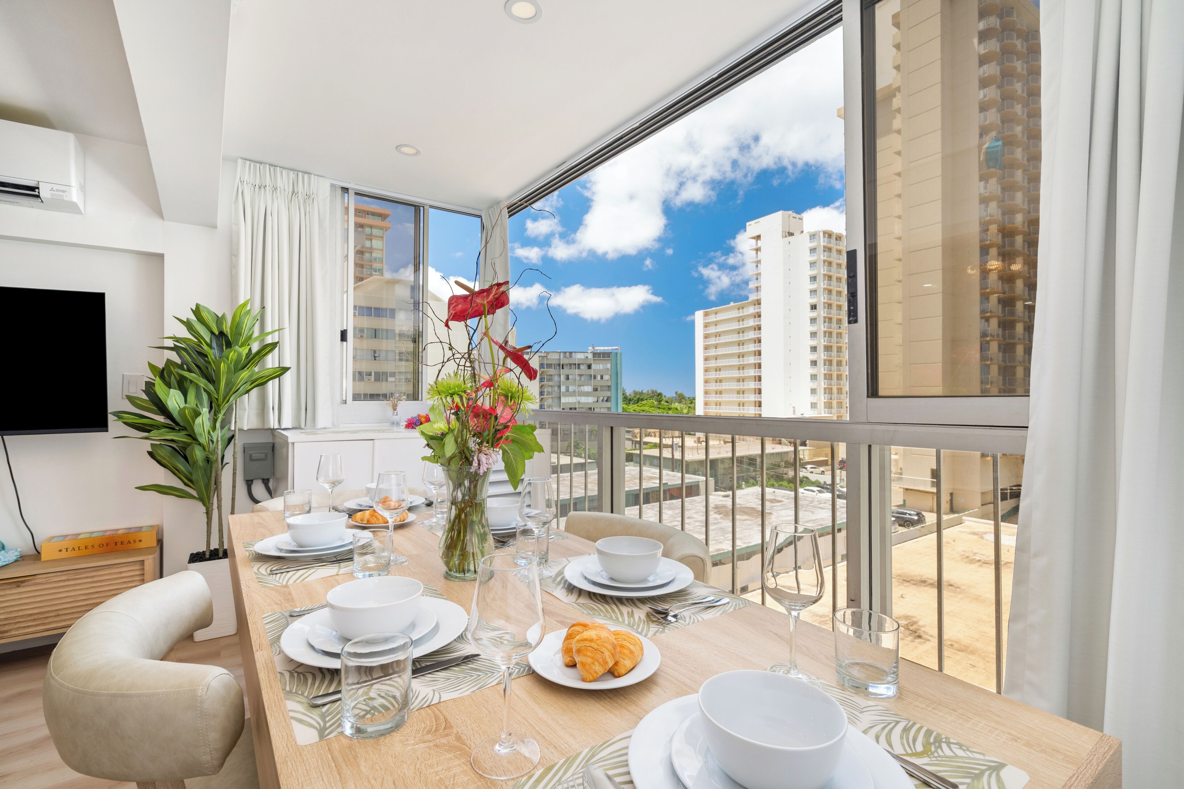 A dining area with a table set for two, a vase with flowers, and a view of the cityscape through large windows.
