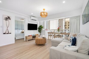 A modern living room with a sofa, a TV, a dining area, and a large window with a view of buildings. - 2-bedroom apartment in serene Honolulu with AC for relaxing stays (Honolulu)