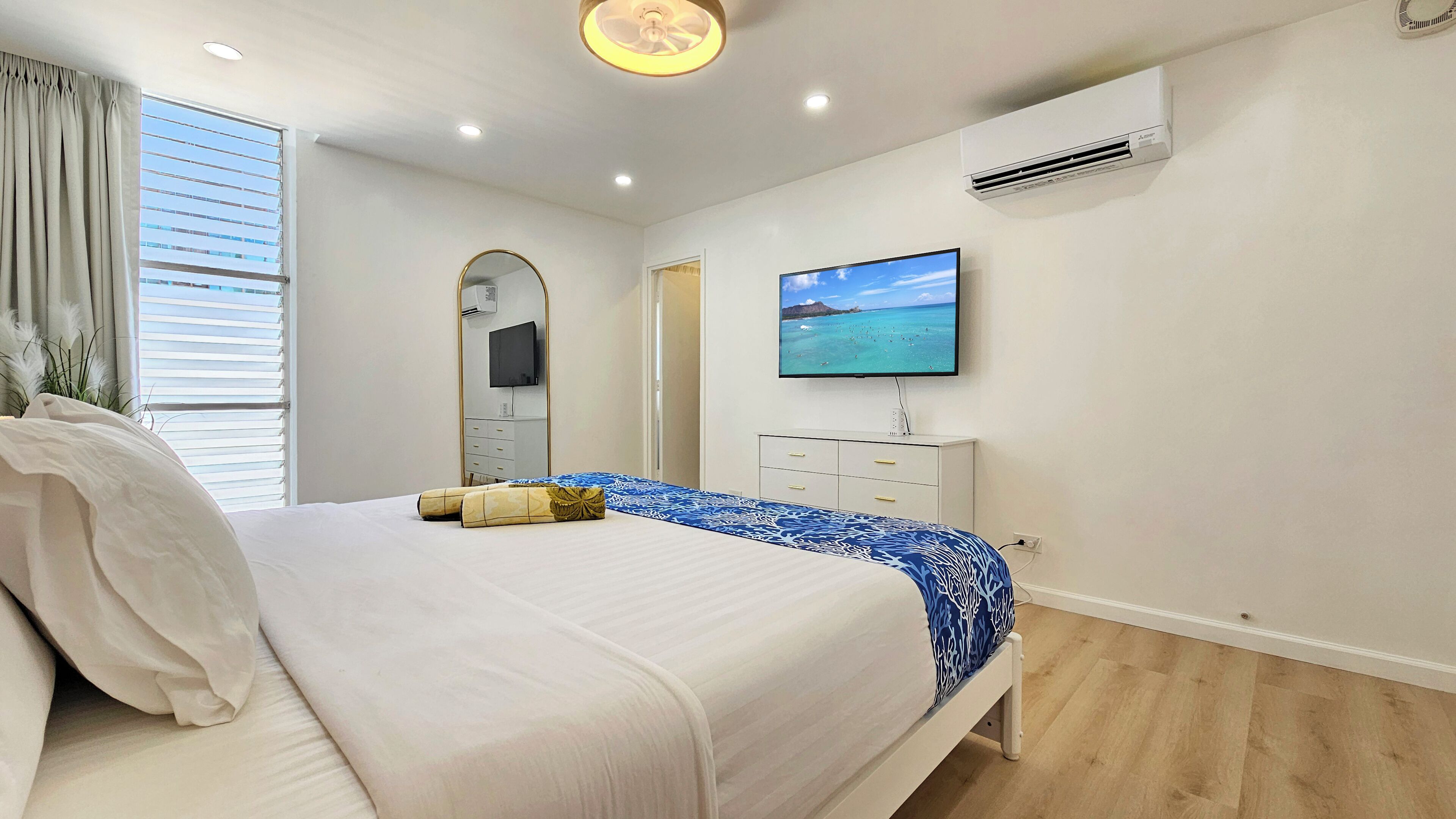 A bedroom with a bed, a television, an air conditioning unit, and a window with blinds.