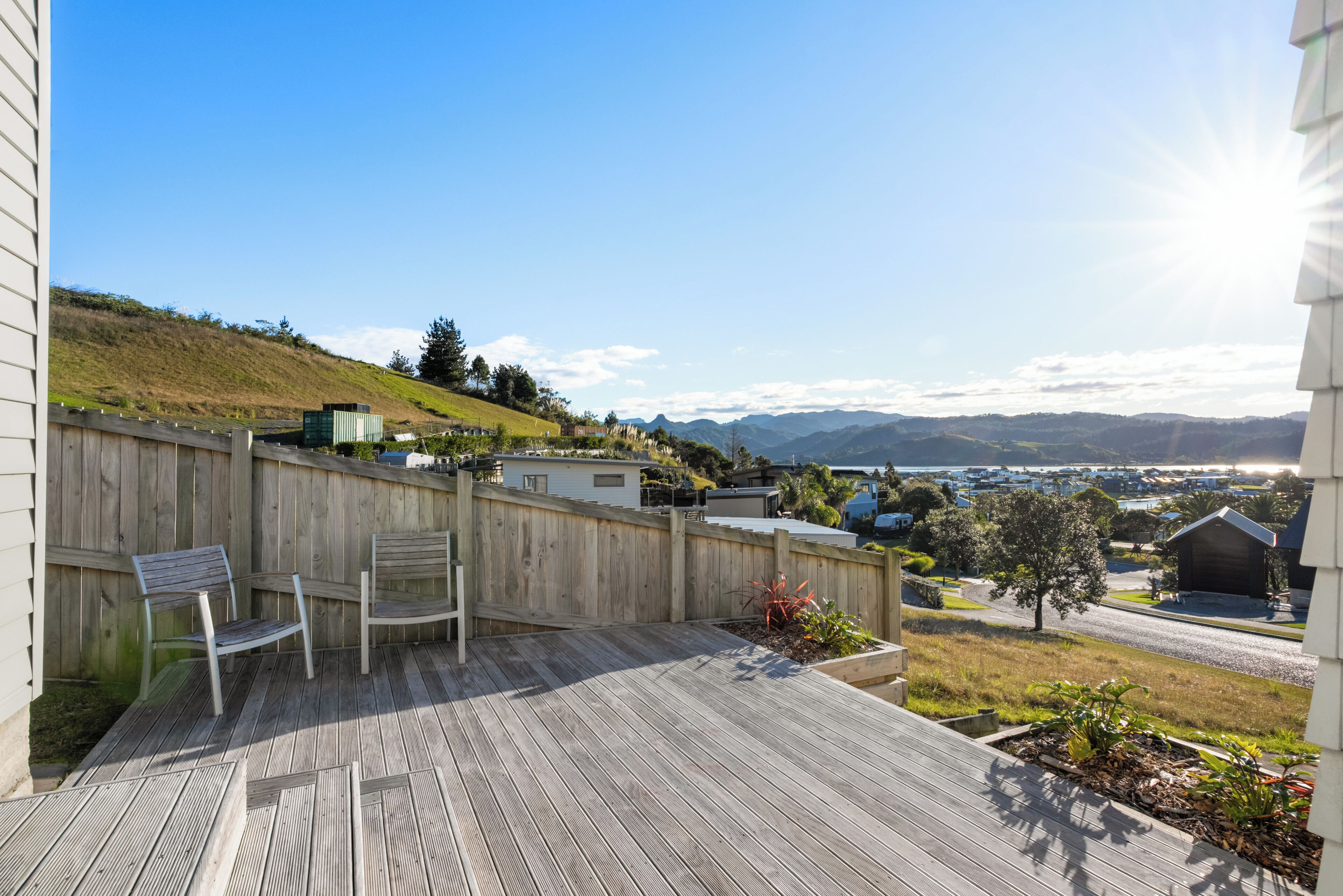 Una terraza de madera con dos sillas, una cerca de madera y vista a un paisaje montañoso con edificios a lo lejos.