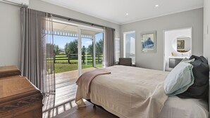 A bedroom with a large bed, a wooden dresser, a bathroom with a sink and mirror, and a sliding glass door leading to a patio. - Meadow Escape - Mangawhai Holiday Home (Mangawhai)
