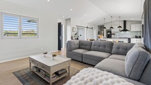 A modern living room with a grey sectional sofa, a white coffee table, and a view of the kitchen in the background. - Meadow Escape - Mangawhai Holiday Home (Mangawhai)