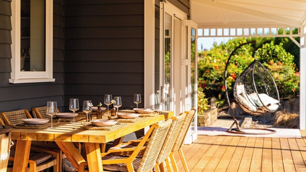 A wooden deck with a table set for dining, outdoor chairs, and a hanging chair. - Meadow Escape - Mangawhai Holiday Home (Mangawhai)