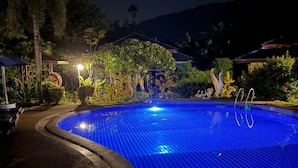 Una piscina nocturna con superficie de azulejos azules, rodeada de follaje tropical y un pabellón.