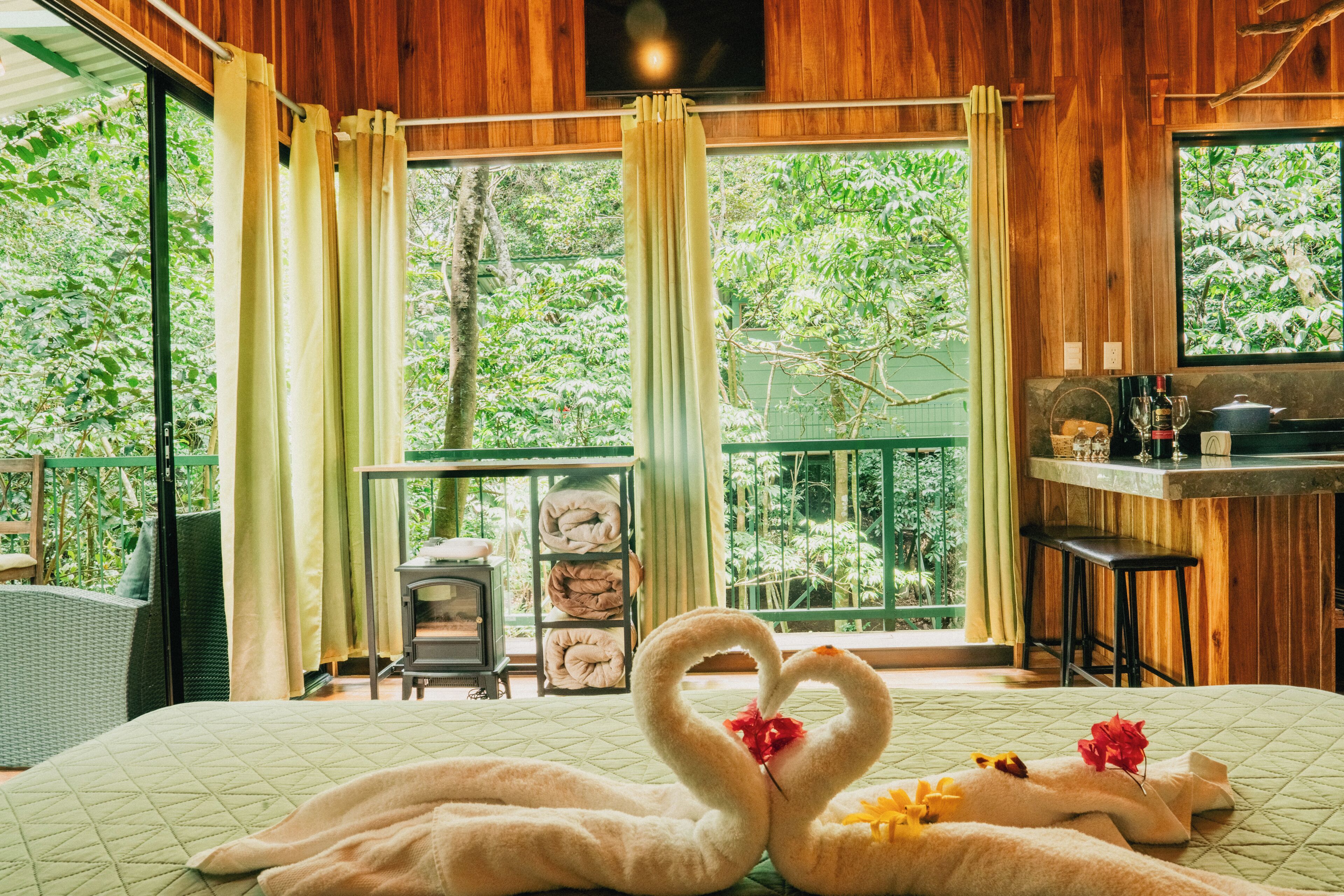 A room with a bed, a heart-shaped swan decoration, a balcony with a view of trees, and a small bar area.