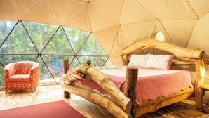 Alojamiento con tienda de campaña, cama de madera y una silla acolchada, con vista al bosque.