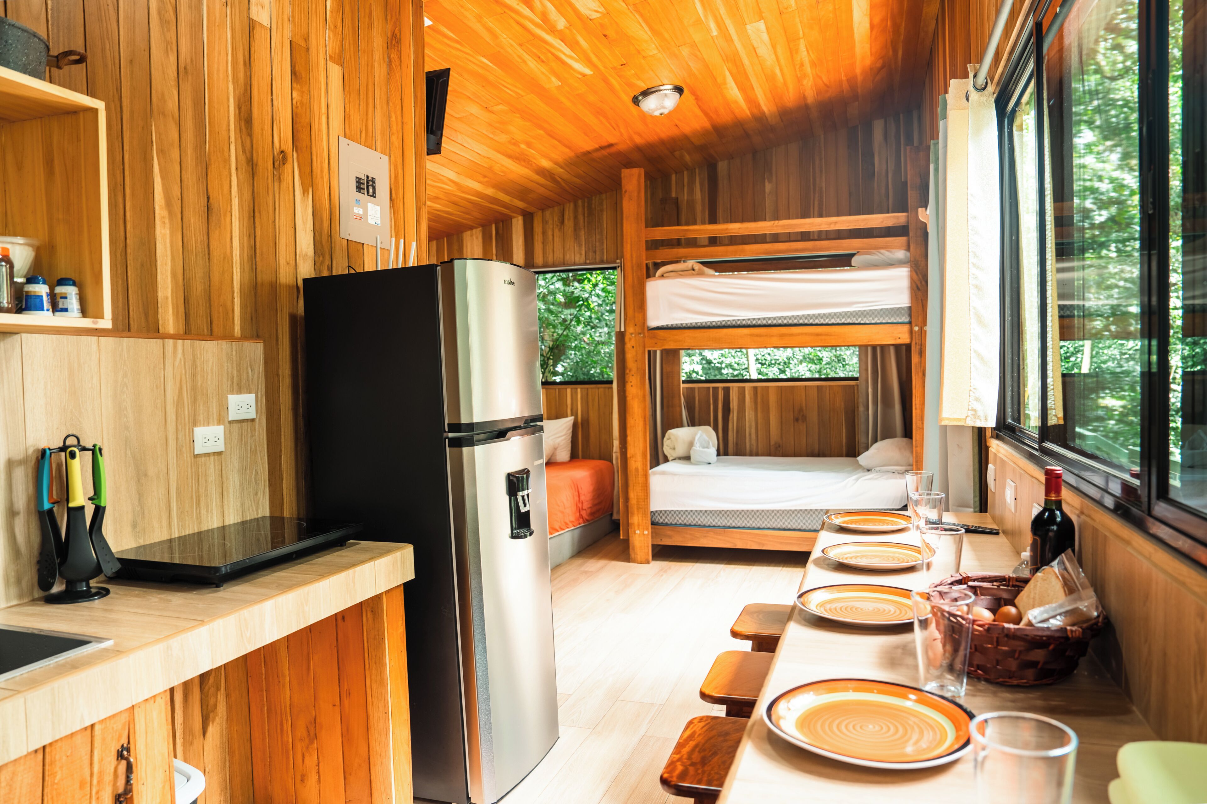 Une cuisine compacte équipée d’un réfrigérateur, de meubles en bois, d’une table à manger avec assiettes et verres, et d’un lit superposé donnant sur une fenêtre.