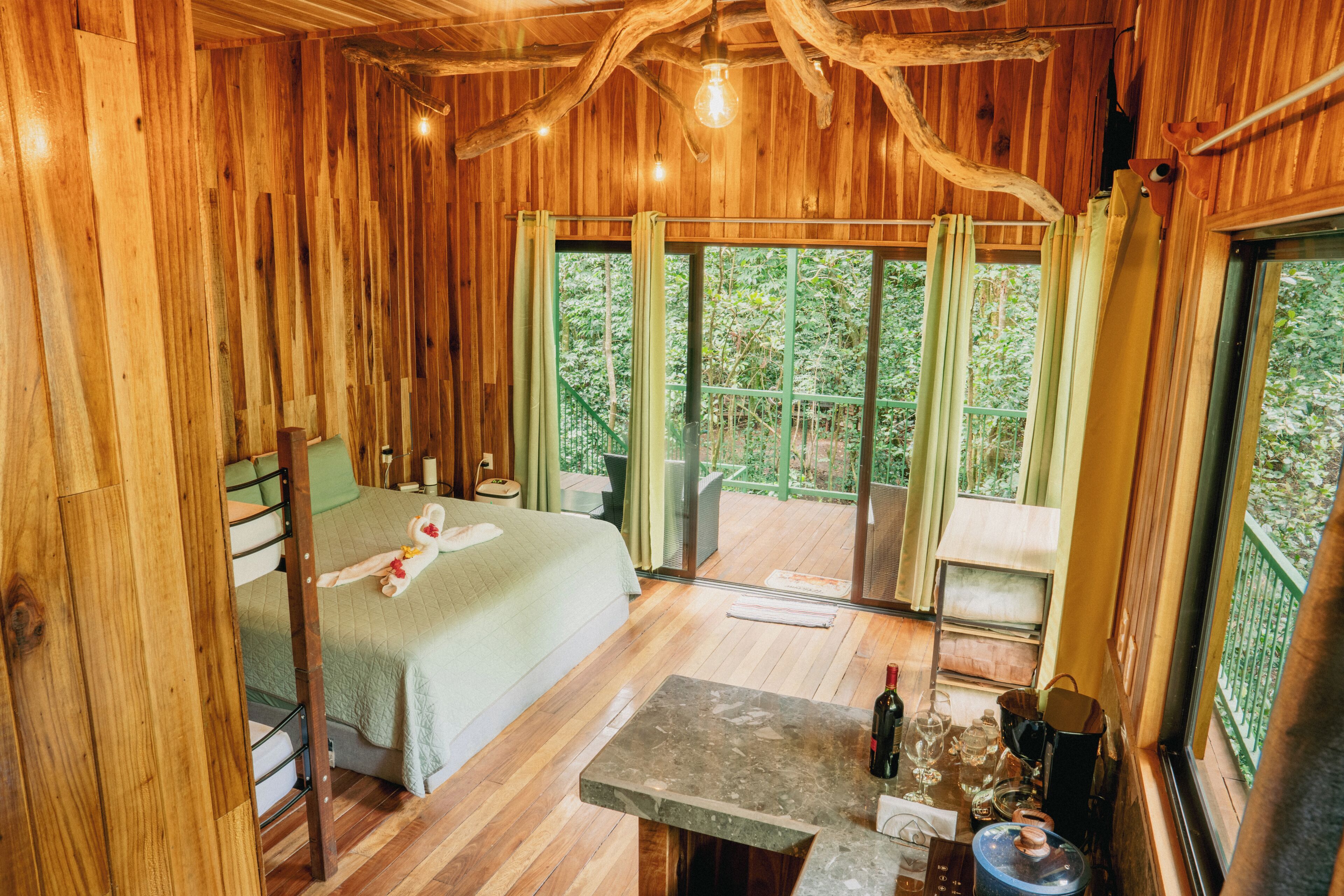 A wooden room with a bed, a table with bottles, and a view of greenery outside.