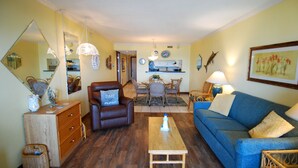 Un salon avec un canapé, une table basse, un fauteuil marron, un coin repas avec une table et des chaises, et une cuisine en arrière-plan.