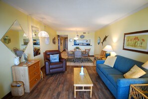 A living room with a sofa, a coffee table, a brown armchair, a dining area with a table and chairs, and a kitchen in the background. - Land's End Resort - Relaxing Oceanfront Getaway with Private Balcony & Resort Am (Myrtle Beach)