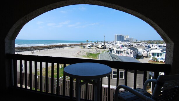 Une vue à travers une fenêtre en arc d’une propriété située en bord de mer, dotée d’un balcon, d’une table et de chaises.