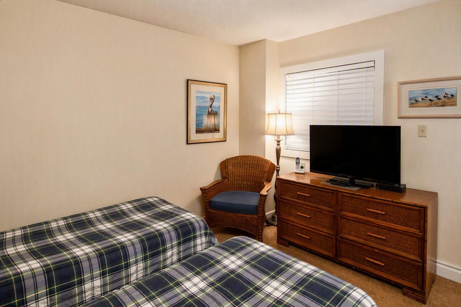 Een hotelkamer met twee bedden met ruitpatroon, een rieten stoel, een houten dressoir met een televisie en een raam met jaloezieën.