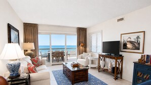 A living room with a television, a coffee table, a sofa, a chair, and a bookshelf. There is a view of the ocean through the sliding glass doors. - Land's End Resort - Oceanfront Views & Resort Amenities (Myrtle Beach)