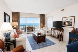 A living room with a television, a coffee table, a sofa, a chair, and a bookshelf. There is a view of the ocean through the sliding glass doors. - Land's End Resort - Oceanfront Views & Resort Amenities (Myrtle Beach)