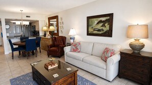 A living room with a sofa, coffee table, and a lamp. There is a dining area with chairs and a table. A kitchen is visible in the background. - Land's End Resort - Oceanfront Views & Resort Amenities (Myrtle Beach)