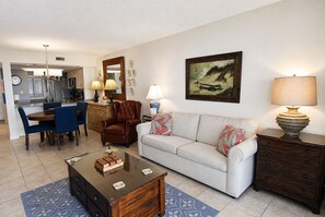 A living room with a sofa, coffee table, and a lamp. There is a dining area with chairs and a table. A kitchen is visible in the background. - Land's End Resort - Oceanfront Views & Resort Amenities (Myrtle Beach)
