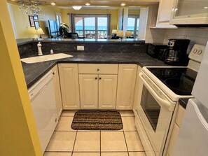 A compact kitchen with a dishwasher, stove, and a view of the ocean through the sliding glass door. - Land's End Resort - Family Retreat with Ocean Views, Pools & Spacious Layout (Myrtle Beach)