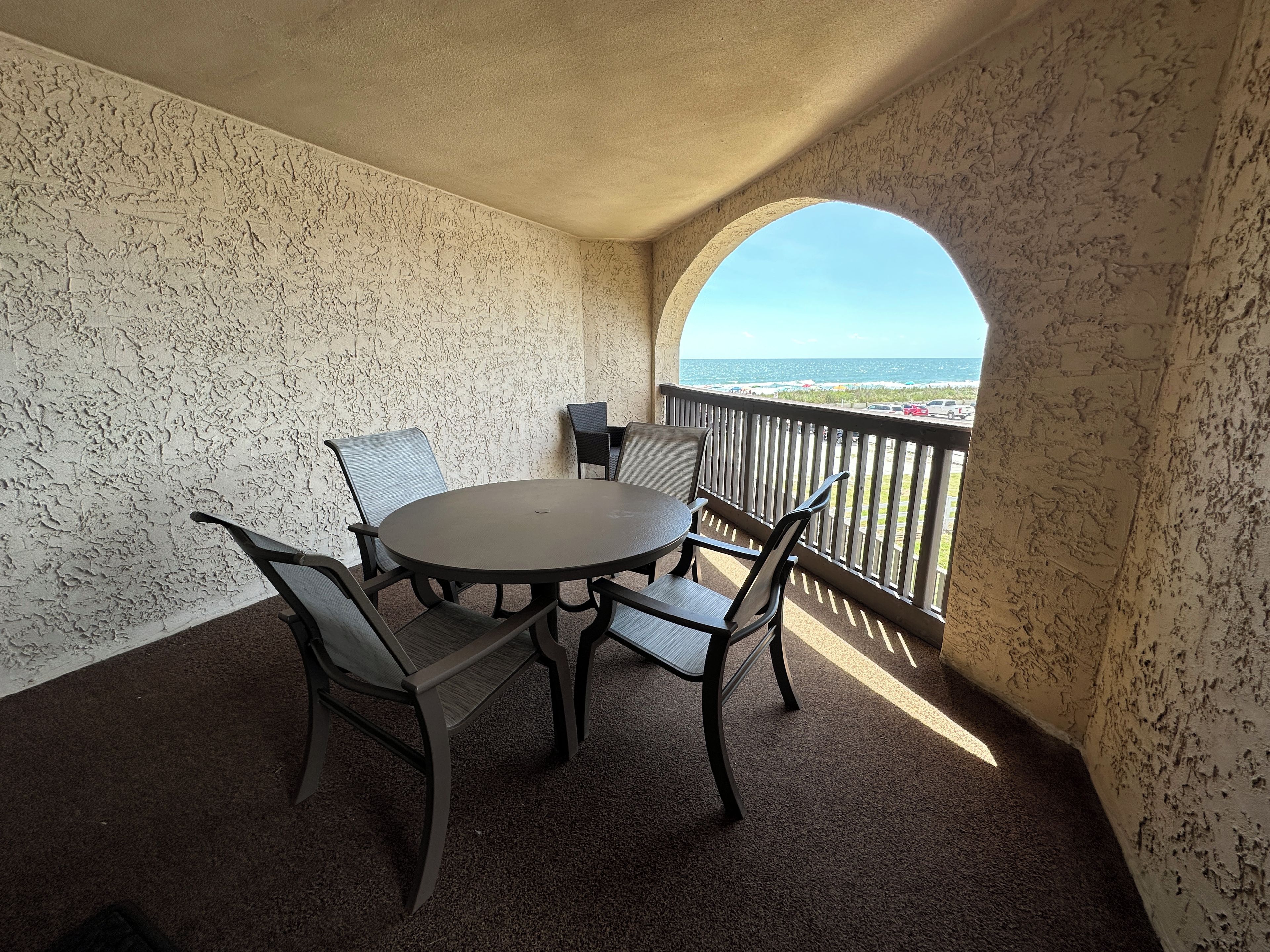 Ein Balkon mit rundem Tisch und vier Stühlen, mit Blick auf einen Strand und einen Parkplatz, wo das Meer klar zu sehen ist.