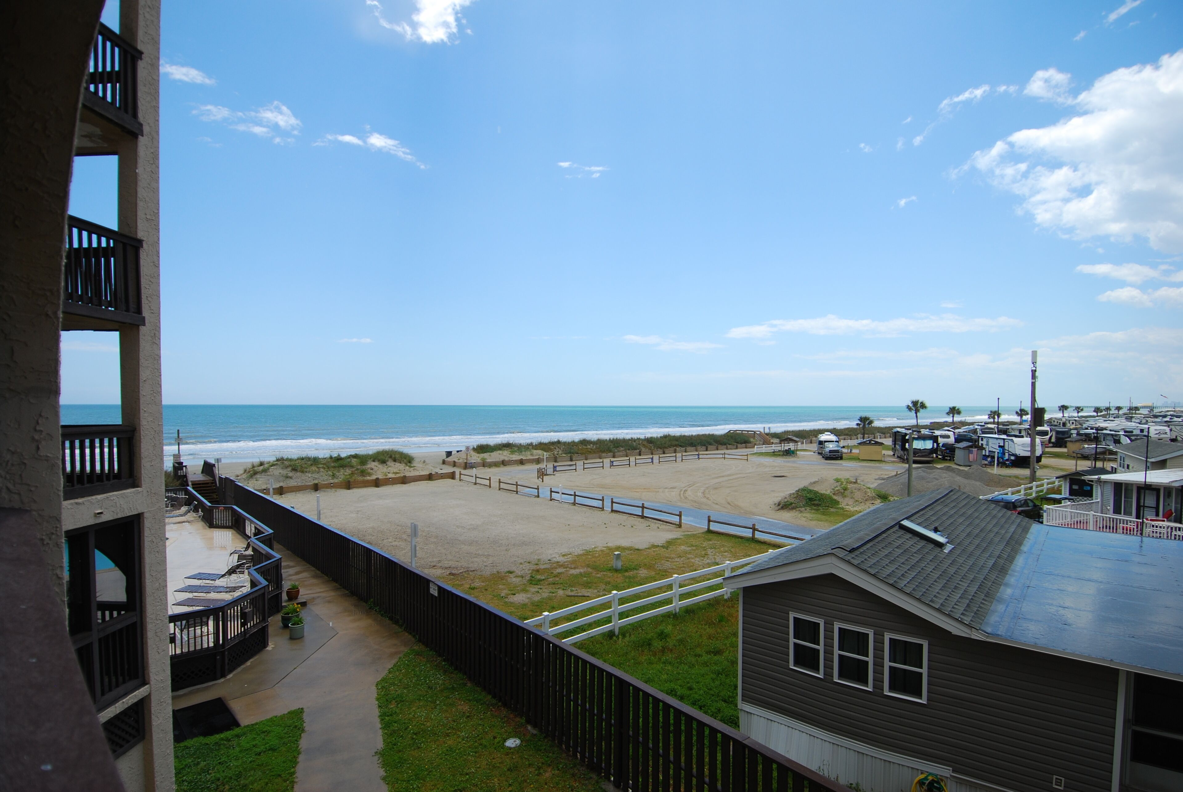 A view of a beach from a balcony with a clear sky, a sandy beach, and a parking area with vehicles.