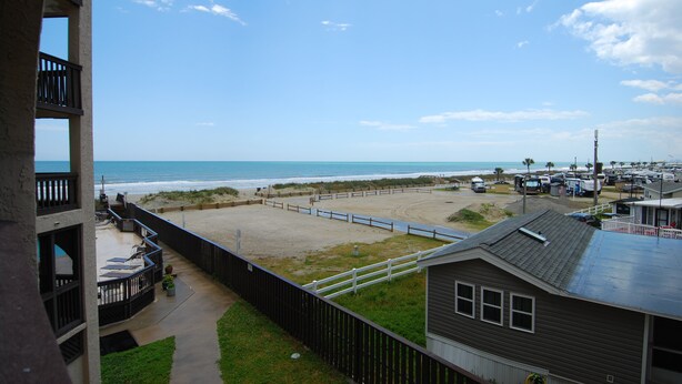 Blick auf einen Strand von einem Balkon aus, mit blauem Himmel, sandigem Ufer und einem Parkplatz mit Fahrzeugen.