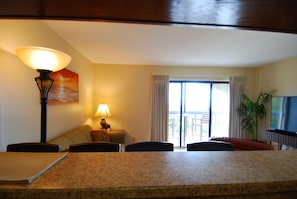A hotel room with a dining area featuring a granite table, chairs, a lamp, a television, and a large window with curtains. - Ocean View | King Suite, Balcony & Beach Access (Myrtle Beach)