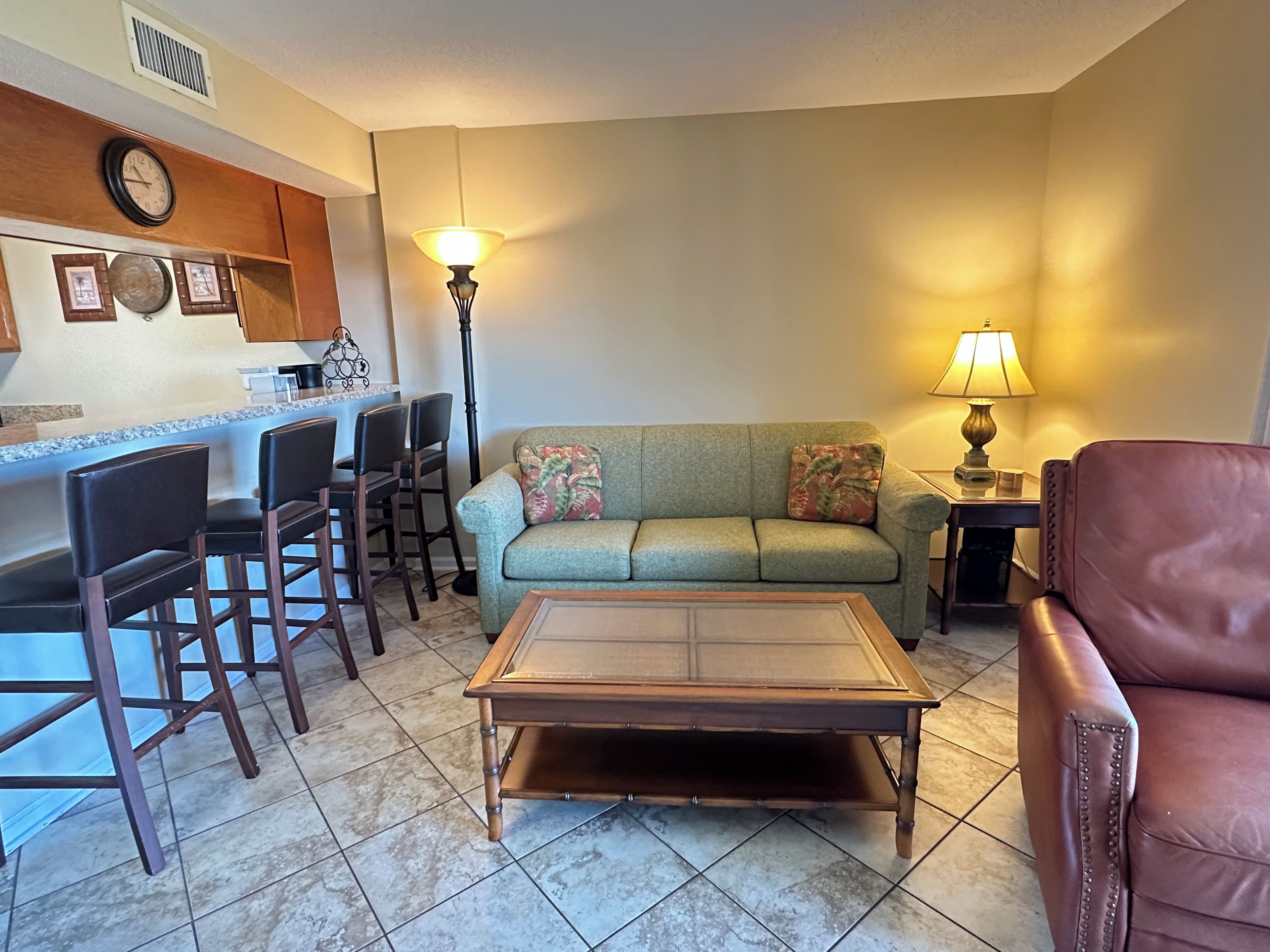 A living room with a sofa, a coffee table, a dining area with chairs, and a kitchen area with a clock and a lamp.