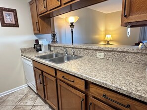 A kitchen with granite countertops, wooden cabinets, a double sink, a coffee maker, and a wall-mounted lamp. - Ocean View | King Suite, Balcony & Beach Access (Myrtle Beach)